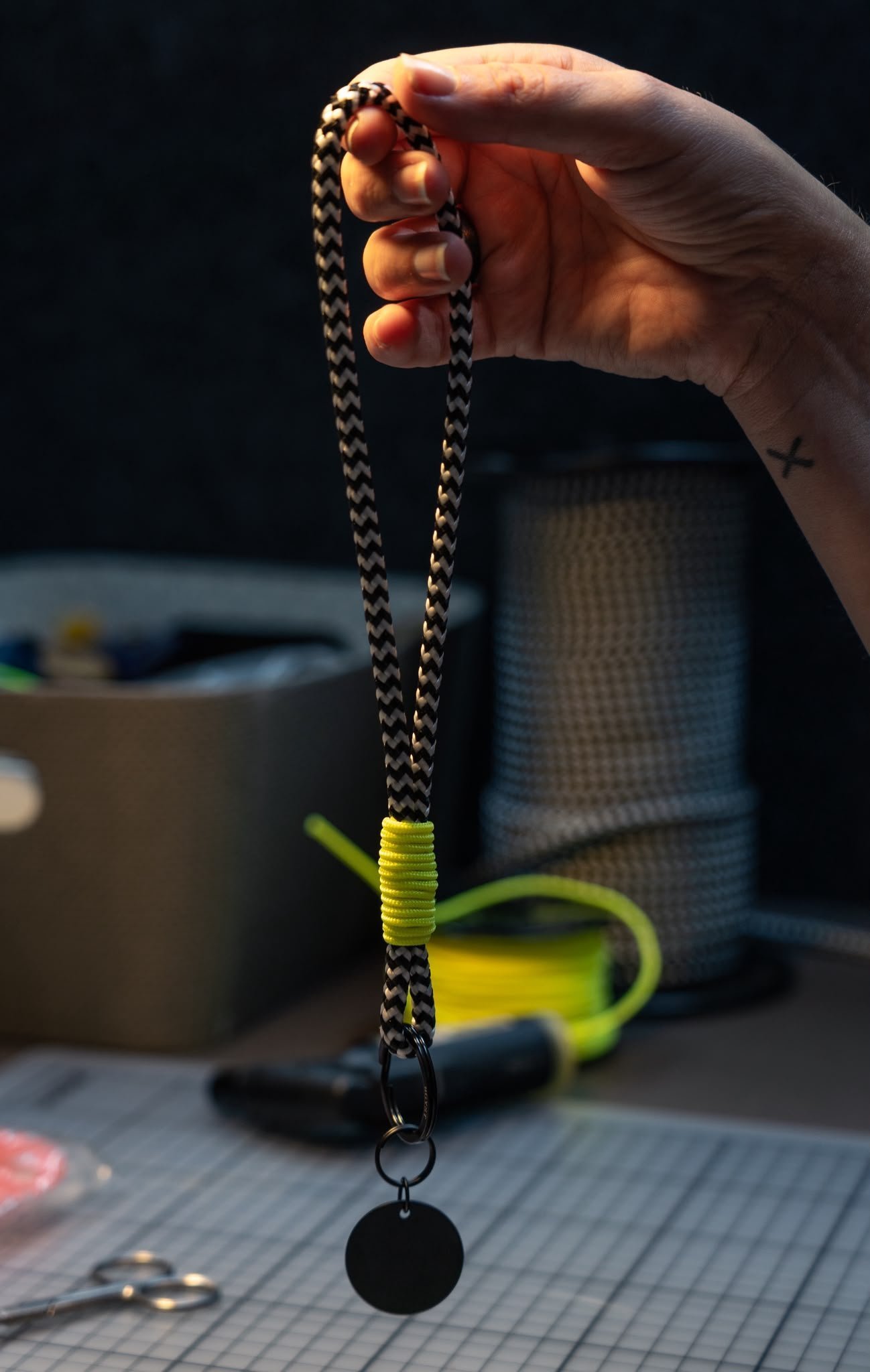 *gyst Schlüsselband mit schwarz-weisser Seilschlaufe, neongelber Wicklung, schwarzen Ringen und rundem schwarzem Anhänger; eine Hand mit X-Tattoo hält es im Zürcher Atelier.